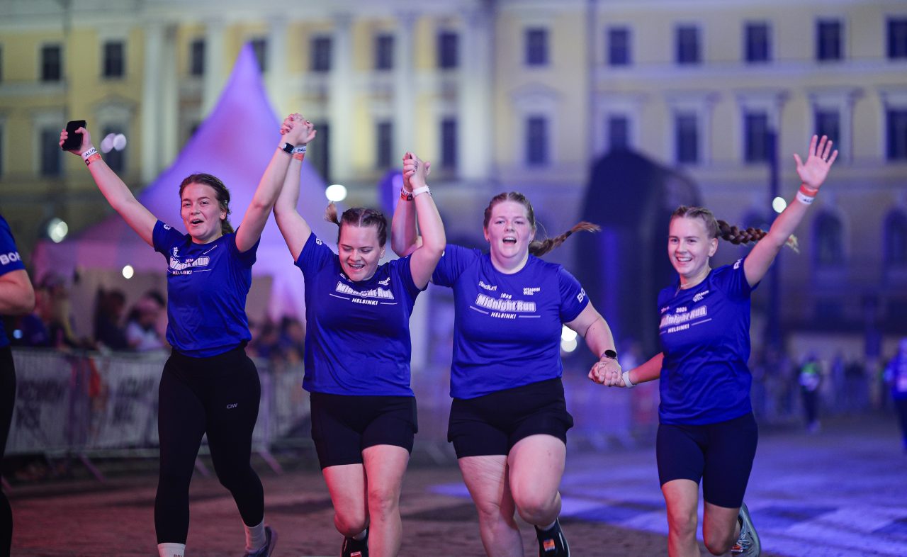 Fyra tjejer springer hand i hand med händerna i luften över mållinjen och firar under Midnight Run Helsinki 2026