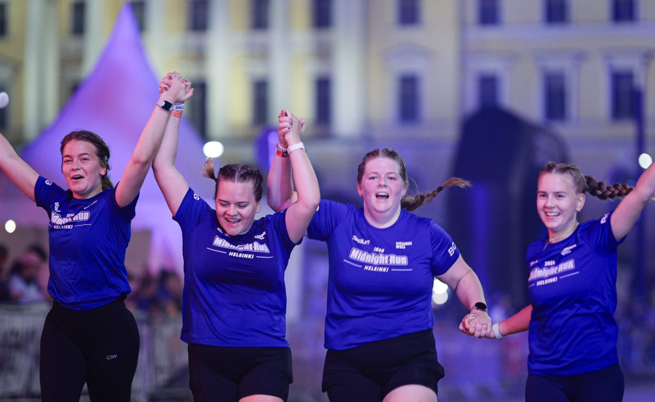 Fyra tjejer springer hand i hand med händerna i luften över mållinjen och firar under Midnight Run Helsinki 2026