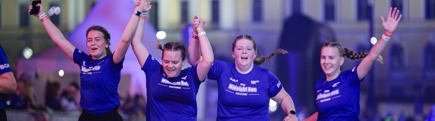 Fyra tjejer springer hand i hand med händerna i luften över mållinjen och firar under Midnight Run Helsinki 2026